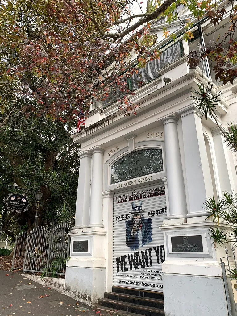 This is a photograph of the former Theosophical Society Hall at 371 Queen Street, Auckland, New Zealand. The building is now known as "The White House Adult Entertainment Centre". The main entrance is a roller door, closed in this photograph, and has a depiction of "Uncle Sam" pointing at the viewer, with the wording "THE WHITE HOUSE. DANCERS. BAR STAFF. WE WANT YOU. 6 DAYS 8PM-6AM" followed by contact phone numbers and the establishment's URL.