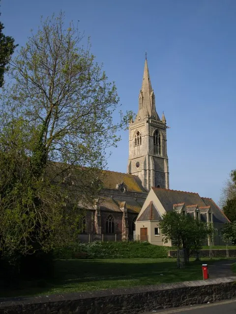 All Saints' church, Babbacombe. Another view of 1302963 from across St Anne's Road. https://historicengland.org.uk/listing/the-list/list-entry/1280043 describes the church in detail, with the tower and spire thus: "4-stage west tower with a stone spire with tall louvred lucarnes; projecting south-east 5-sided stair turret; set back buttresses rise to belfry stage."