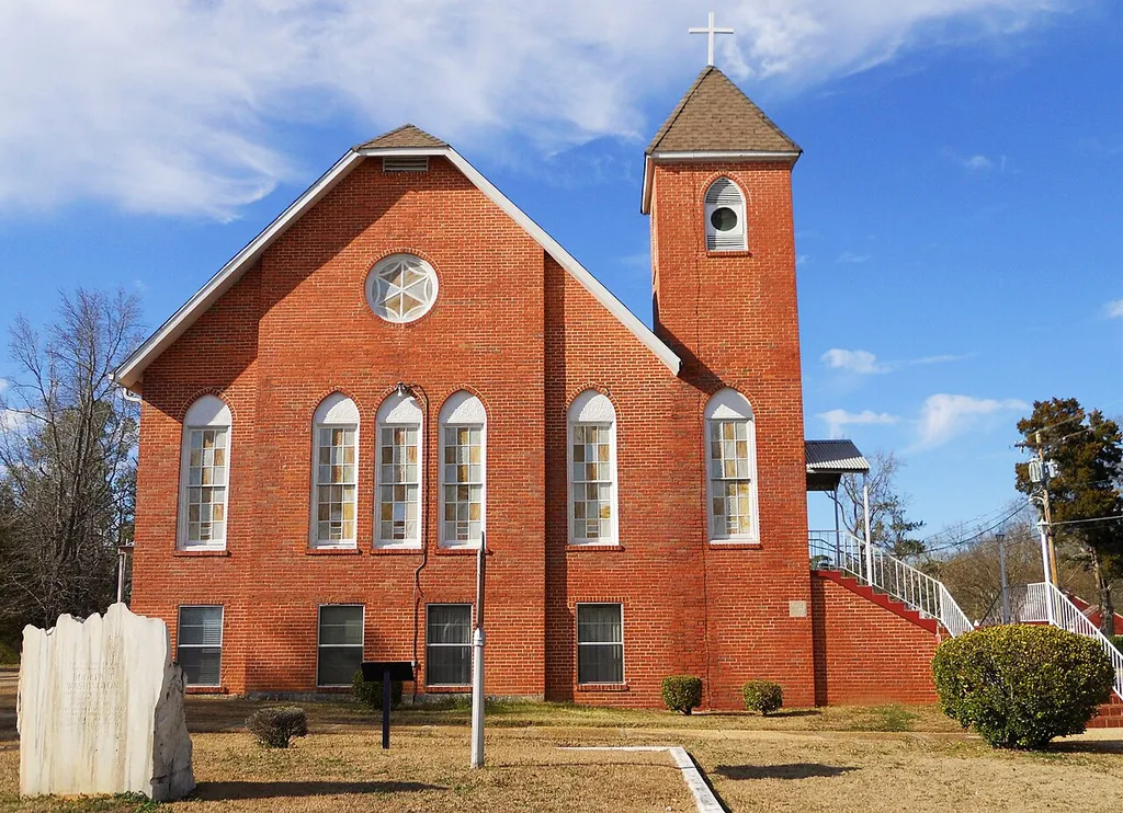 This is a photograph of Butler Chapel African Methodist Episcopal Zion Church located in Tuskegee, Alabama. "This church was the focal point for a multi-year grass-roots project that united and empowered African Americans, rural and urban, educated and uneducated, to fight for the right to vote. Butler Chapel AME Zion Church, an imposing brick building located on a hill west of downtown Tuskegee, is a prominent landmark in the historically black neighborhood known as Zion Hill. The building, the second church on this site, was constructed about 1877. Originally built in wood, the church was sided with brick in the 1940s. In a 1957 effort to minimize the number of black voters in Tuskegee, Alabama's municipal elections, the state legislature simply redrew the town's political districts, placing Tuskegee Institute and all but a small fraction of black residents outside city limits. To protest this action, Tuskegee's middle-class black community and Macon County's poor black citizens joined forces in a seven-year "Crusade for Citizenship." On June 25, 1957, 3,000 area black residents showed up at Butler Chapel for the first of many weekly mass meetings. Only 500 attendees could fit into the church's small sanctuary; the rest listened outside. Charles Gomillion, a professor at Tuskegee Institute and the driving force of the black Tuskegee Civic Association, urged the crowd to join a "Trade with Friends" boycott of local white merchants. "We are going to buy goods and services from those who help us, from those who make no effort to hinder us, from those who recognize us as first-class citizens," he promised. The boycott ended in early 1961 when city boundaries were returned to their original position, after the Supreme Court ruled that a legislature could not single out an isolated segment of a racial minority for discriminatory treatment." [Source: NPS.gov]