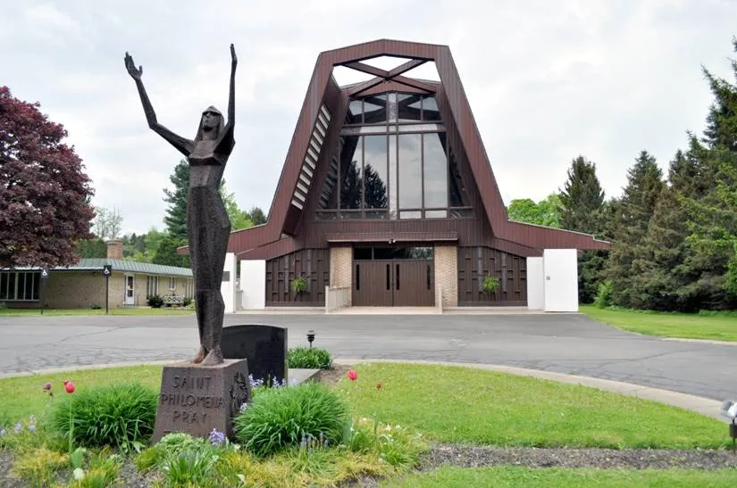 St. Philomena Roman Catholic Church, 26 Plymouth Avenue, Franklinville, New York, July 2011. A striking Modernist church building designed by Buffalo-based architect Gerard Zimmermann (whose Southern Tier work also prominently includes St. Joseph's Catholic Church in Holland), the architecture adapts the A-frame design contemporarily popular in residential architecture for ecclesiastical use, where the steel-frame roof swoops in dramatic geometrical forms from the gable atop the main façade (which frames an enormous recessed stained-glass window where the shape of a cross is delimited by redwood slats in a narrow rectangular pattern echoed on the walls flanking the entrance) to the sides, where the exposed ends of the steel beams overhang the concrete piers that make up the exterior wall. The stained glass, Stations of the Cross, and status of St. Philomena in front of the building are all the work of Buffalo-area sculptor Larry Griffis. St. Philomena's got its start as a parish in 1873 and worshiped for many years at a site adjacent to the Free Methodist Church on South Main Street. The move to the present site, overseen by its then-pastor Rev. Henry J. Romanowski (1920-1985), culminated with the new church's dedication in May 1965. Father Romanowski died in 1985 and is buried on the church grounds. The more recent history of the parish includes its 2007 merger with the former Holy Family R.C. Church in Machias as well as its "link" with Holy Name of Mary parish in Ellicottville (with whom they share a priest, currently Rev. F. Patrick Melfi, and financial resources), both as part of the Buffalo Diocese's "Journey in Faith and Grace" parish consolidation program.